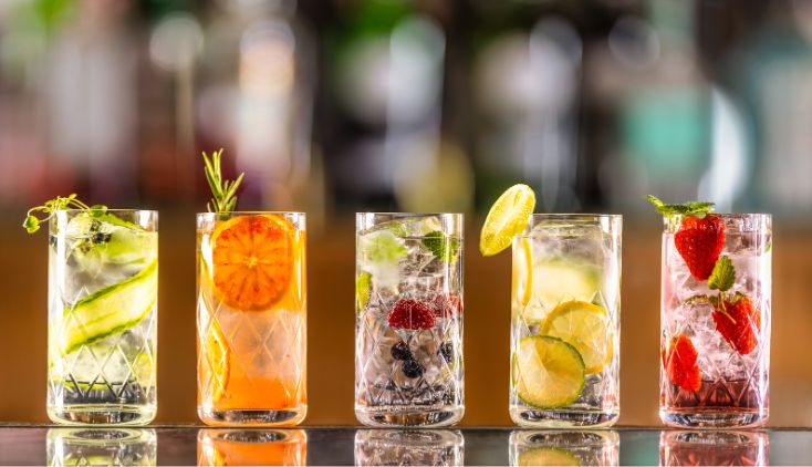 A row of different cocktails in tall glasses, with different fruit inside each one.